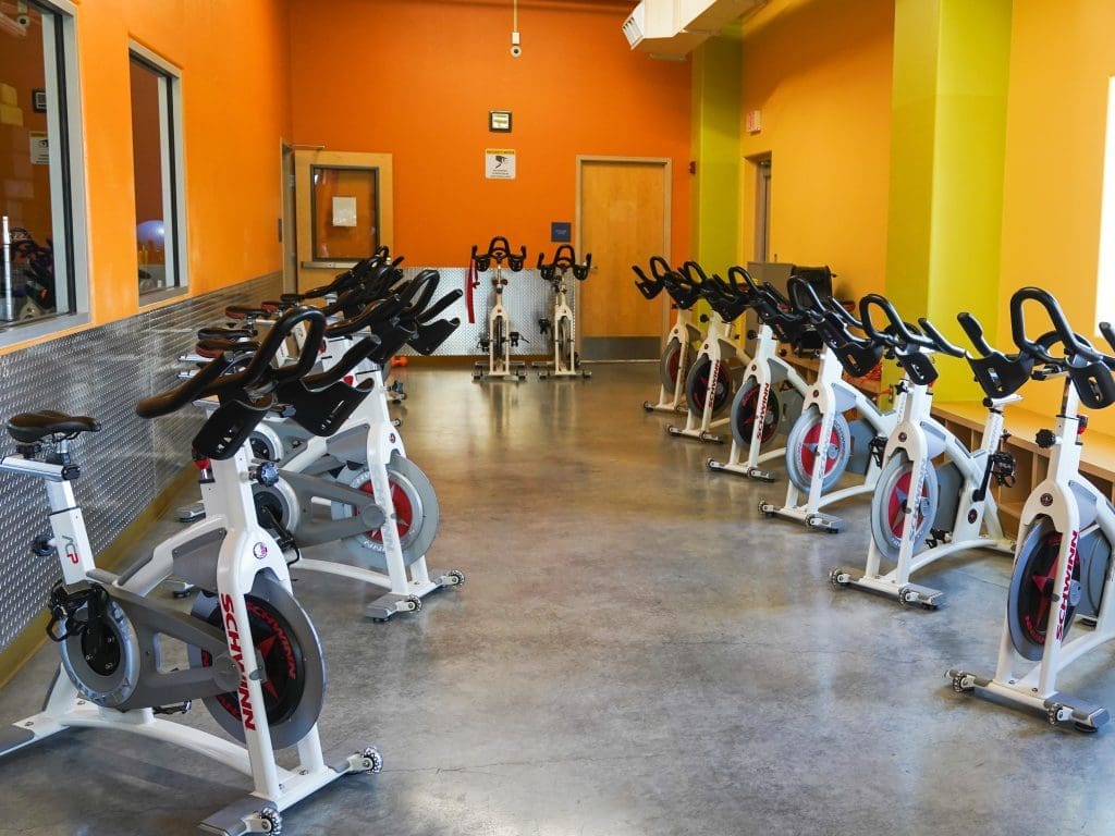 A brightly-lit indoor cycling room with multiple stationary bikes arranged in rows along the walls and center of the room, featuring yellow and orange walls.