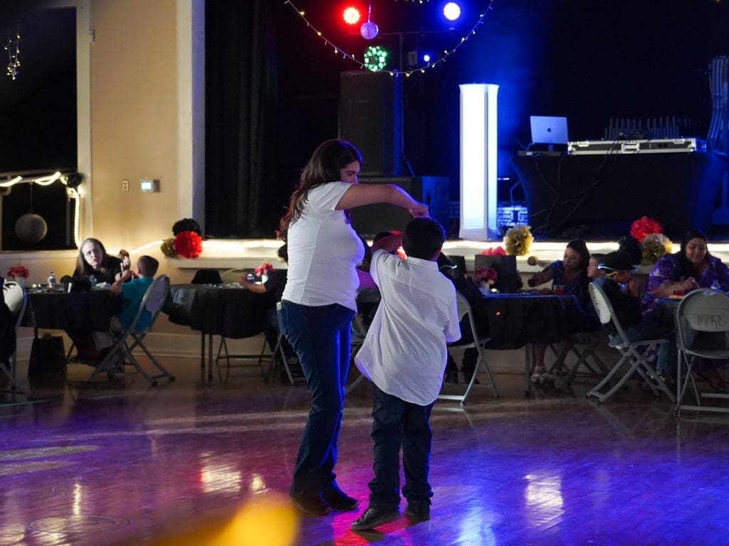 A woman and a child dance together in a dimly lit event hall, surrounded by seated people at tables decorated with flowers. Colored lights illuminate the background.