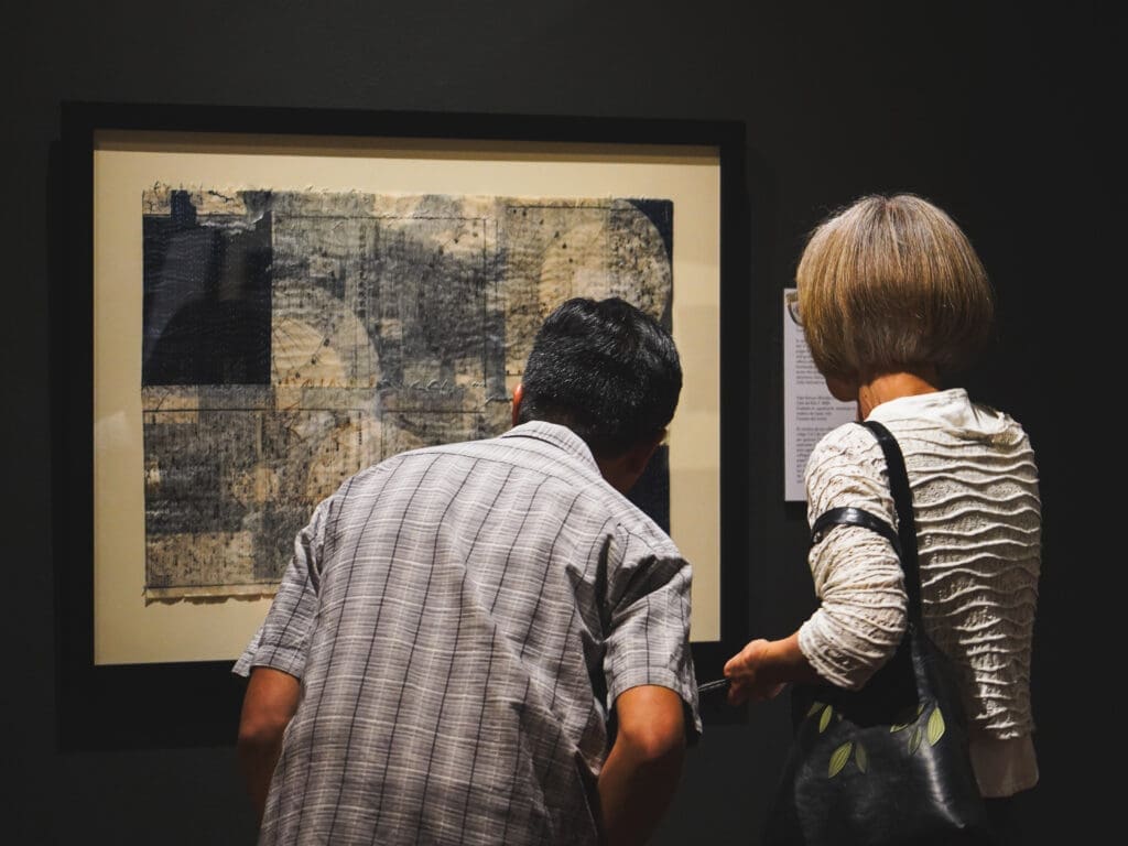 A picture of two people facing away from the camera looking at a blue and white abstract art piece on a dark wall.