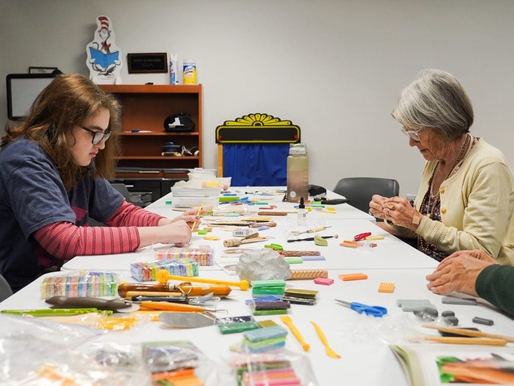 A table full of art and crafts tools. Two people sit on either ends of the table molding clay.