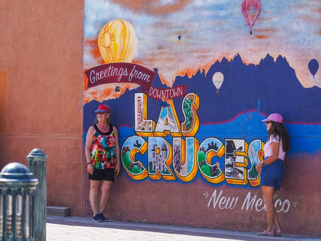 Two women stand in front of a mural of Las Cruces' Organ Mountains with hot air balloons. The mural says "Greetings from the Downtown Las Cruces New Mexico".