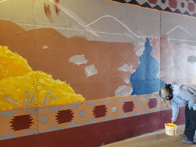 Photo of crew member bent over paint pail dipping brush with mural in background