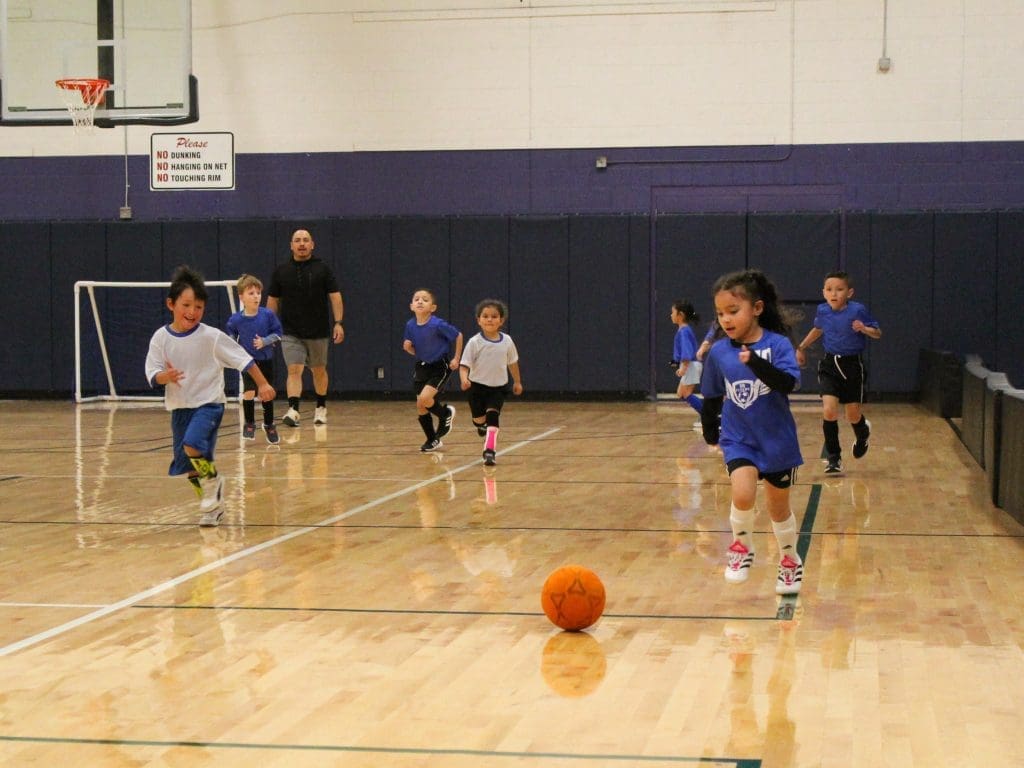 Children playing soccer in an indoor basketball gymnasium.