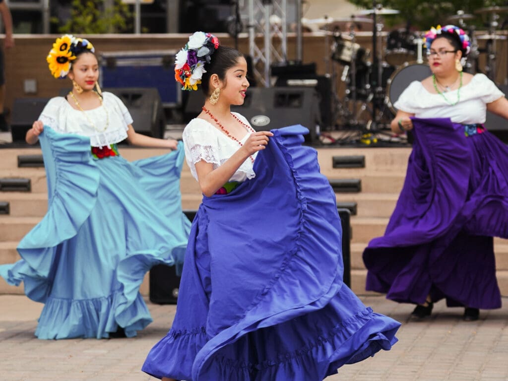 Three dancers in traditional attire, with colorful skirts and flowers in their hair, perform outdoors.