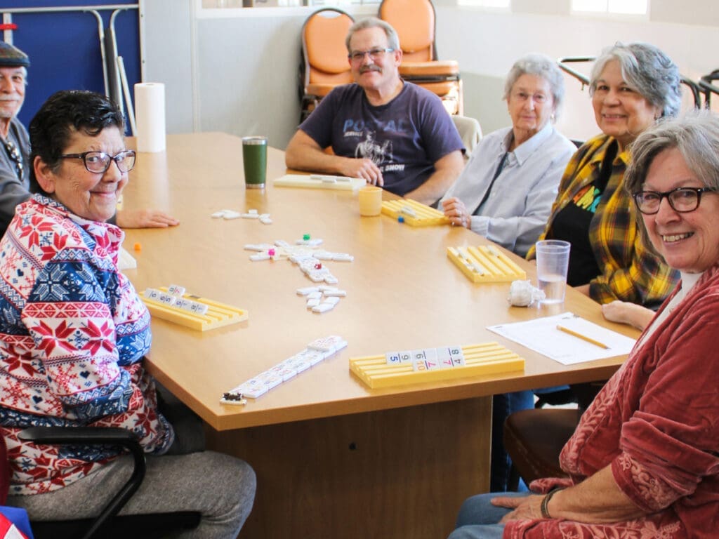 A group of seniors smile at the camera while playing dominoes at a table.