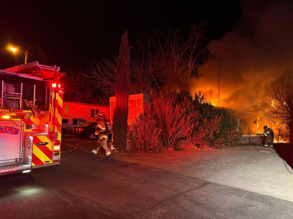 Firefighters fighting a fire at a mobile home with trees and bushes in front of it.