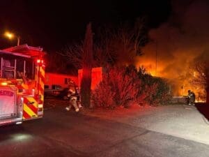 Firefighters fighting a fire at a mobile home with trees and bushes in front of it.