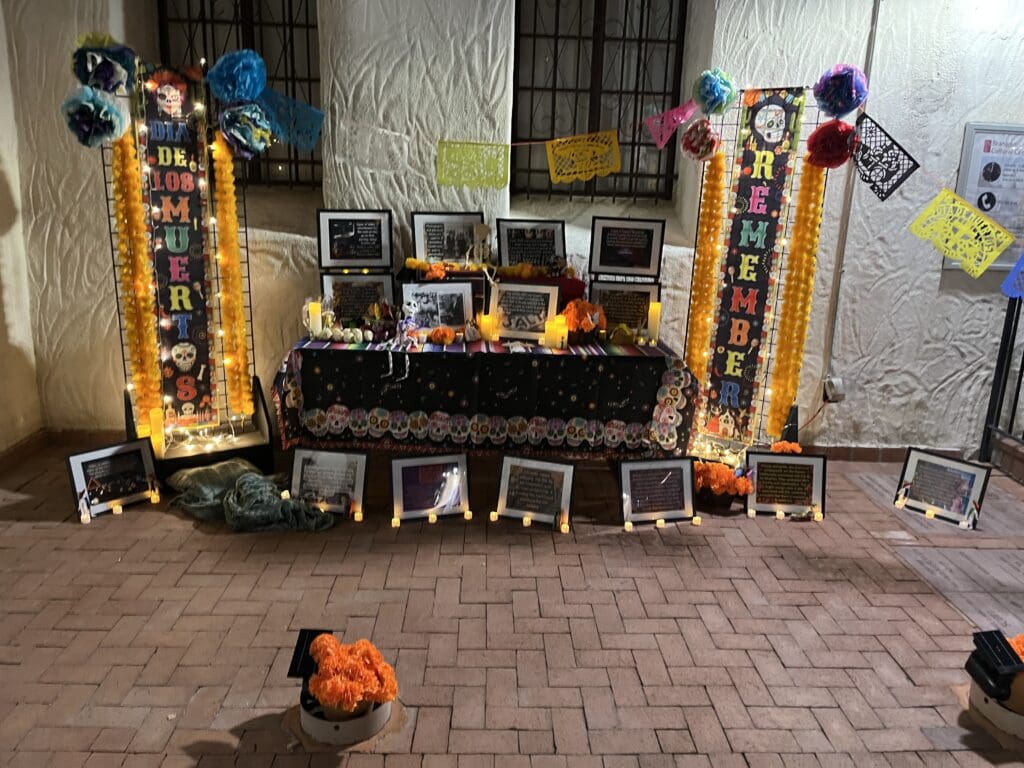 A picture of an ofrenda with a large banner on each side. It has several picture frames filled with information on it and is lit up with lights.