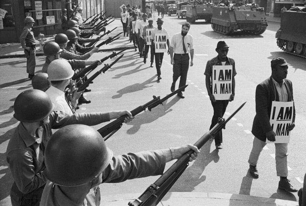 A photo of men walking at Civil Rights Protest wearing banners that say "I AM A MAN" in bold text. Soldiers aim guns at them.
