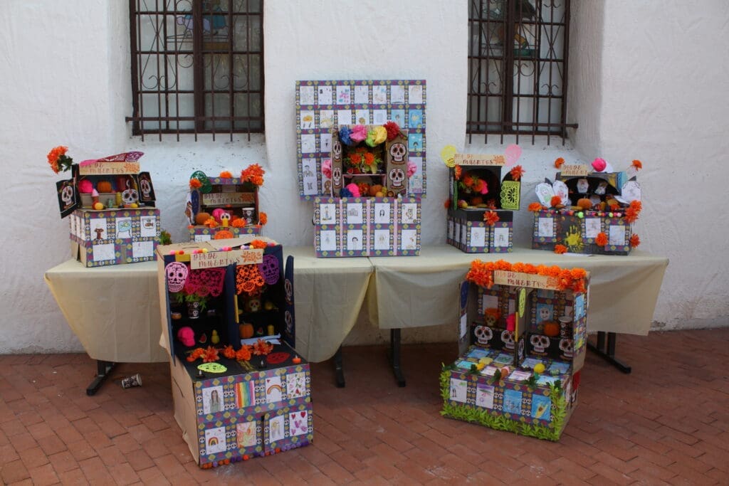 A picture of an ofrenda with several carboard miniature ofrendas on top of it with two in front as well. Each is decorated wth drawings, skulls, and marigolds.