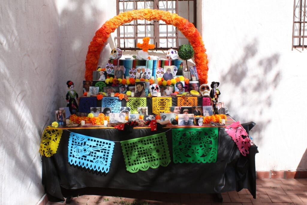 A community ofrenda with an arch of marigolds over it. It is three tiered with papel picado banners, skulls, candles, and pictures.