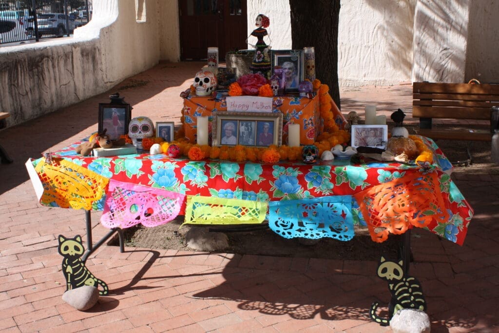 A picture of a two tiered ofrenda in front of a tree with pictures, candles, skulls, and papel picado banners.