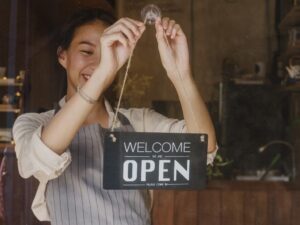 Woman holding up a "Welcome - Open" sign at a front door of a business