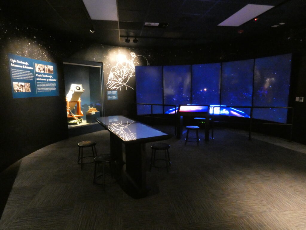 A picture of the Tombaugh room at the Museu of Nature & Science. It is dark with only the screens on the walls, the Stellar Playground interactive table, and the Grazer Gazer telescope behind glass lluminated.