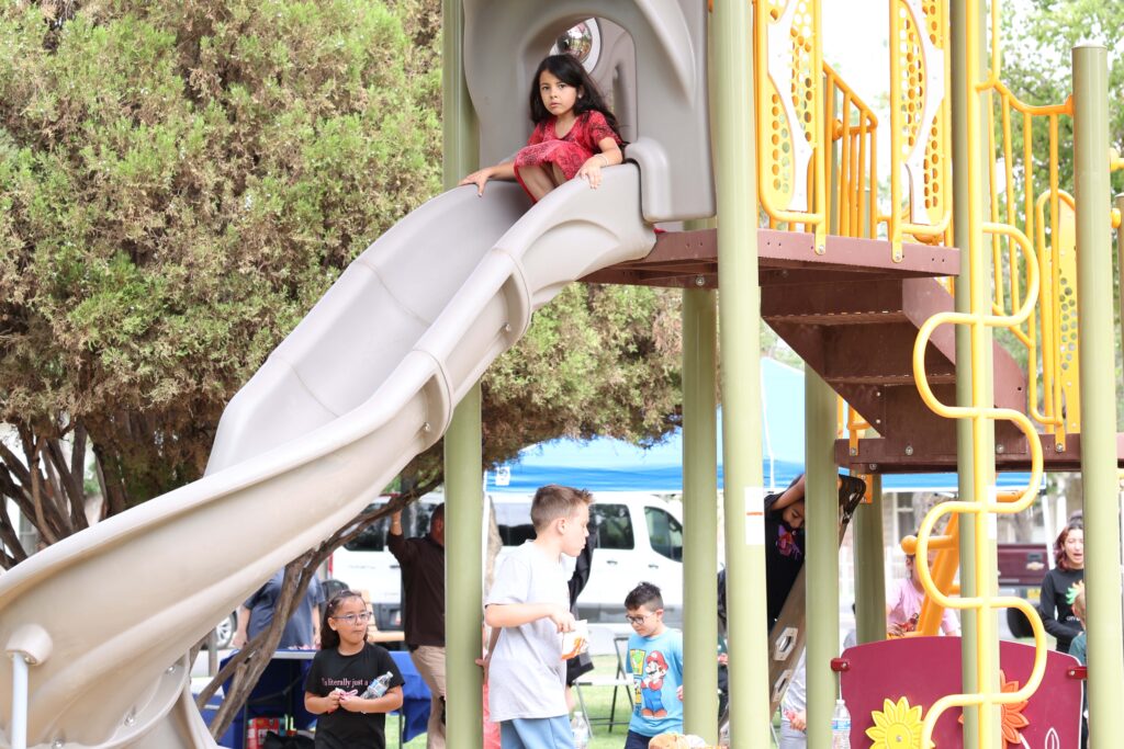 Girl on slide looking at camera while other kids play beneath slide at a park.