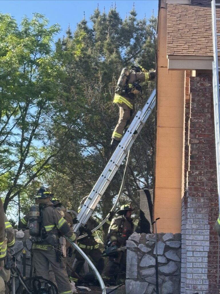 Group of firefighters on the ground while one firefighter is climbing a ladder to the second story of a house.
