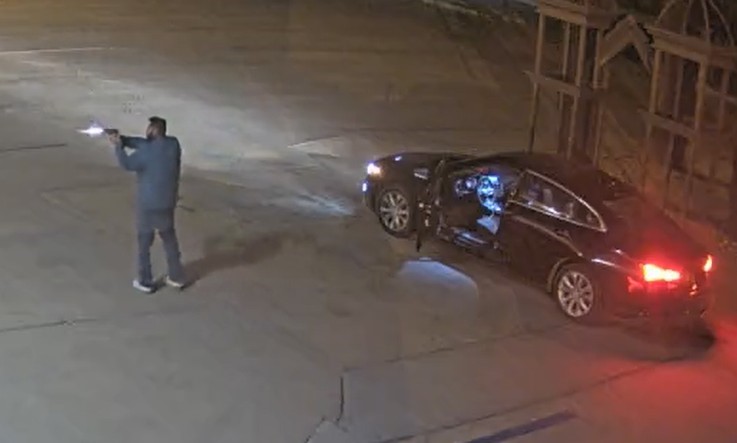 Man shooting a gun in a parking lot next to a car with the driver's door opened.