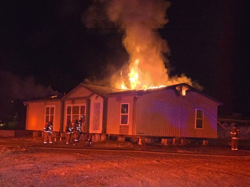 Firefighters standing outside a house with flames coming out of the roof at night.