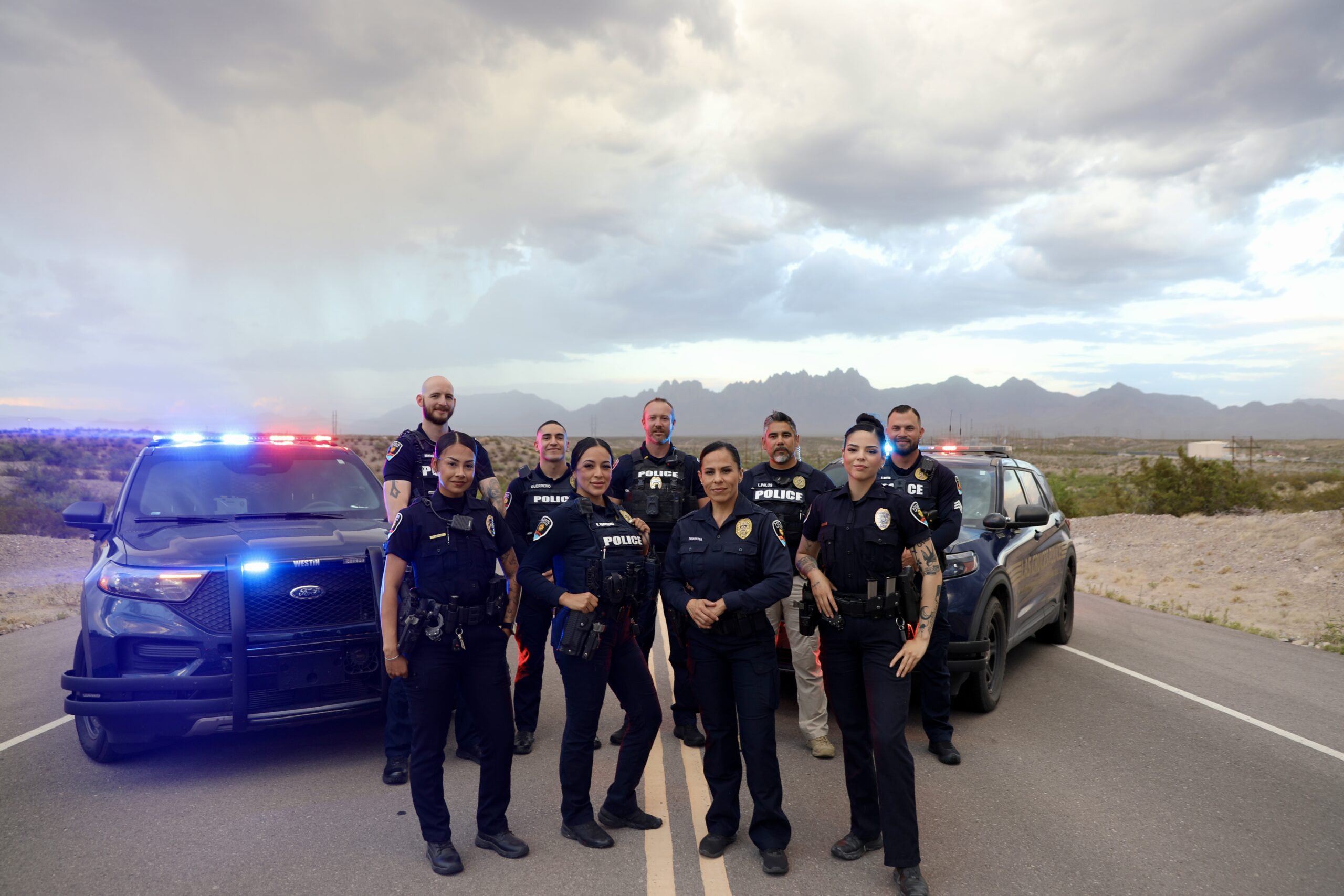 Nine (9) LCPD officers standing in front of two cruisers with their red and blue lights on.