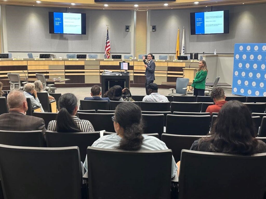 A man and a woman standing in front of an audience in City Council chambers.