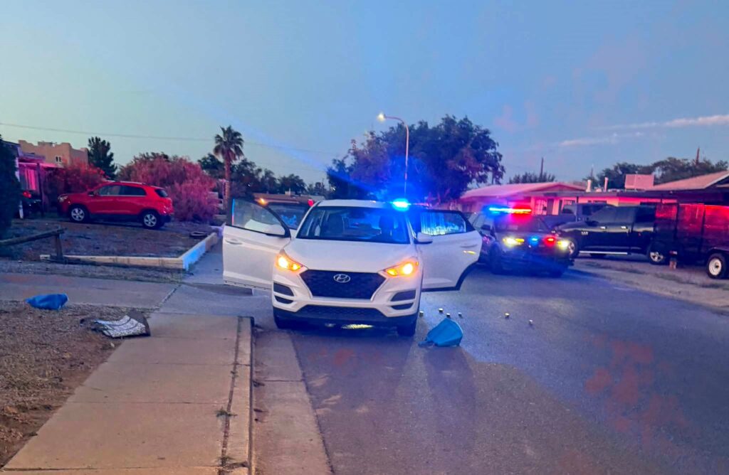A white sedan in the road with its doors open and a police vehicle with its lights on parked in the road also.
