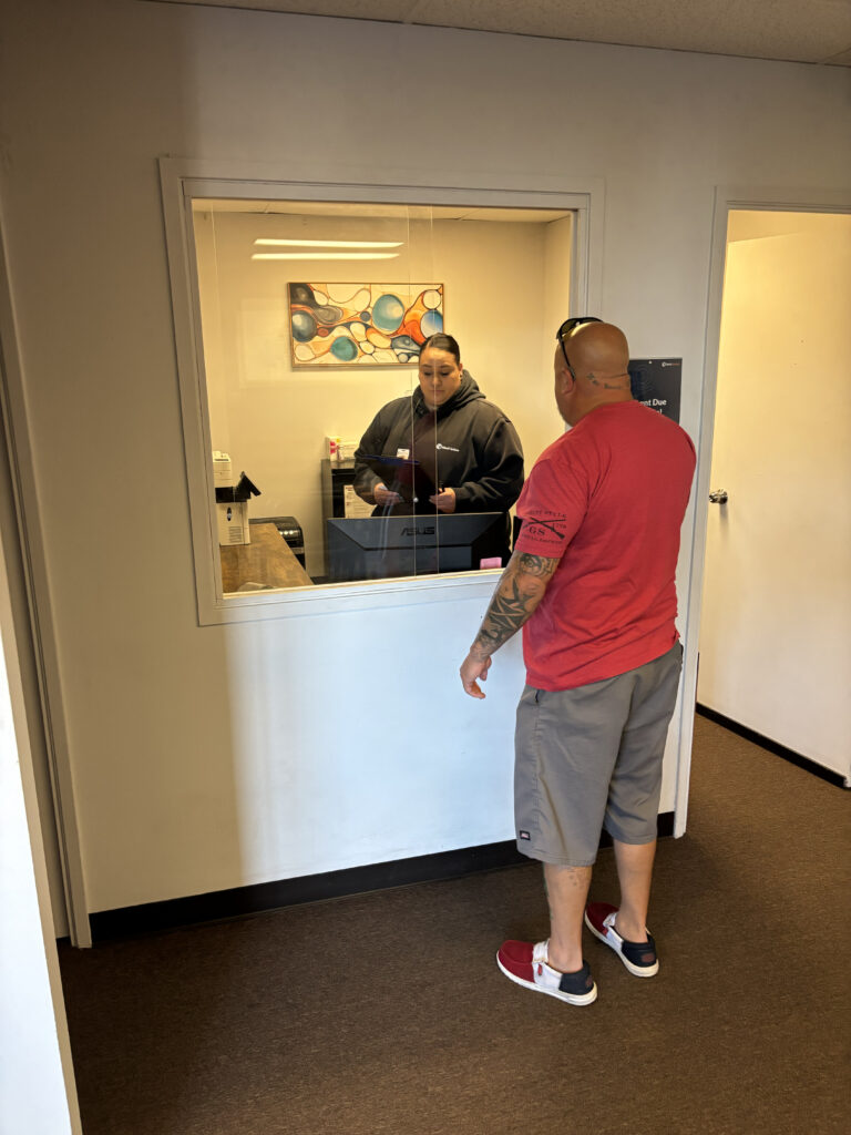 Man standing at a service window talking to a woman with a clipboard on the other side of the window.