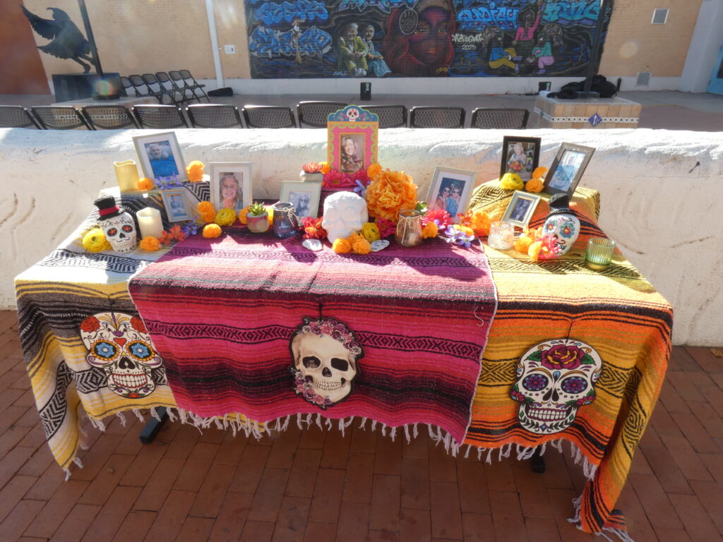 A community ofrenda with an arch of marigolds over it. It is three tiered with papel picado banners, skulls, candles, and pictures.