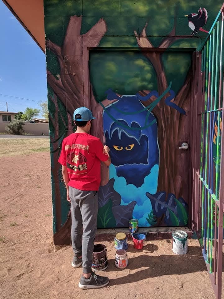 Teen male wearing red tshirt and blue cap painting mural on wall.