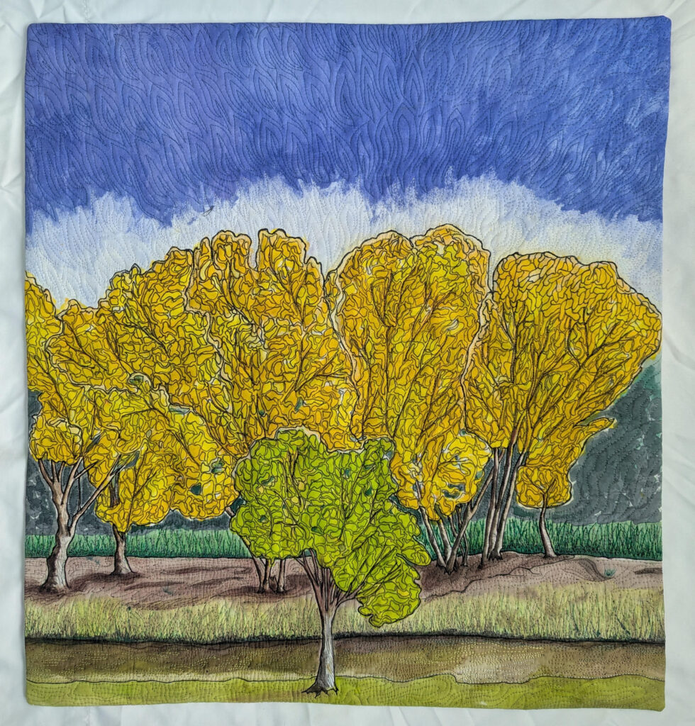 Quilted artwork featuring a row of yellow trees, one green tree in the foreground, grassy ground, and a blue and white sky above.