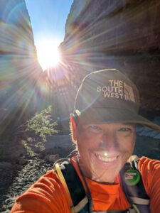Woman wearing cap and backpack against a backdrop of a canyon with sun streaming above the canyon top.