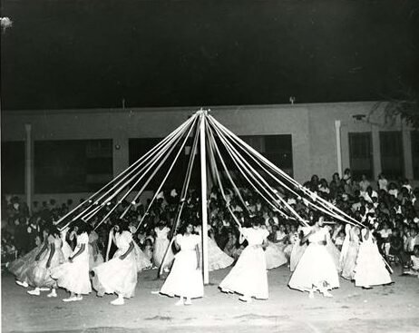 A black and white photo of young girls in matching dresses dancing around a maypole holding ribbons.