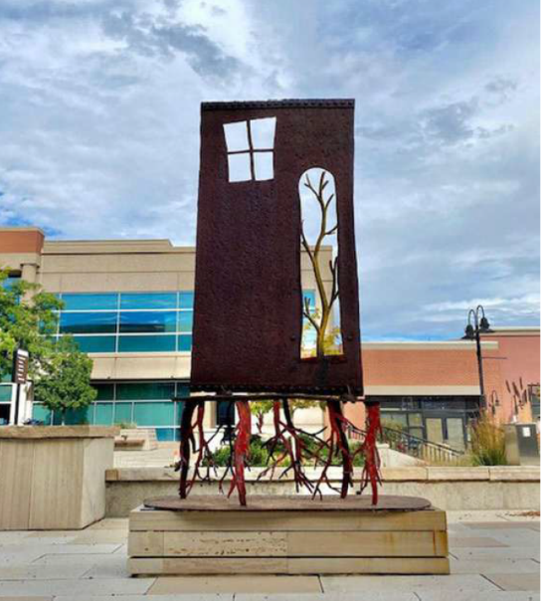 Sculpture that looks like a door with a small window frame on the top left and an open area with a tree silhouette. The base is rootlike steel legs.