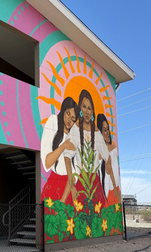 Mural on side of building of three women standing close with plants in front of them.