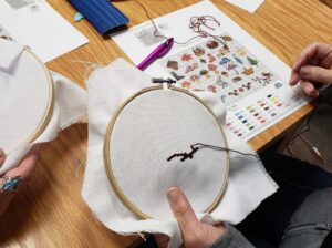 Person sitting at a table cross stitching.
